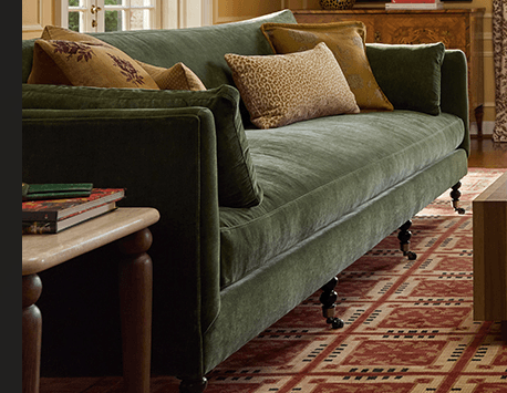 A traditional three-seater sofa upholstered in a rich, deep olive green velvet fabric. It features straight track arms, plush loose seat and back cushions, and stands on dark-finished turned wooden legs equipped with brass casters. Several decorative throw pillows are placed on the sofa, adding to its classic aesthetic. — 3D model