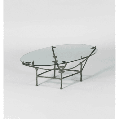 An oval coffee table with a clear glass top supported by a sculptural metal base. The base appears to be cast bronze or a similar metal, with an organic, somewhat branching structure that mimics natural forms. It features four legs that connect to a lower frame, supporting the tabletop via four points. — 3D model (AI enhanced source)