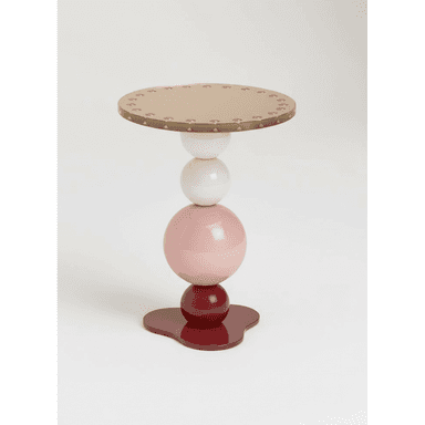 A postmodern accent table featuring a distinctive stacked spherical base in various colors, topped with a circular metallic surface. The base consists of a dark red organic-shaped foot, supporting a smaller dark red sphere, a larger light pink sphere, and a white sphere, arranged vertically. The top is a flat, round surface with a metallic gold or brass finish, detailed with a repeated pattern of small embossed or debossed elements around its edge. — 3D model (AI enhanced source)