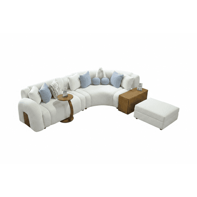 A large, modular sectional sofa featuring a curvilinear design, upholstered in a textured off-white bouclé fabric. It consists of multiple individual seating segments that create a curved arrangement. The set includes an integrated round wooden side table, a rectangular wooden end piece that serves as a surface or storage, and a separate matching upholstered ottoman. Several accent pillows in light blue and a geometric pattern are placed on the sofa. — 3D model (AI enhanced source)
