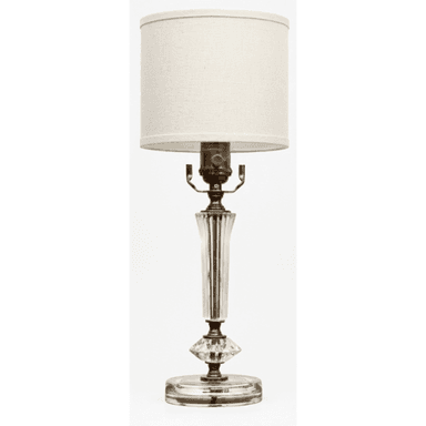 A traditional table lamp featuring a clear glass body with fluted and faceted details, set on a round glass base. It is accented with brass-toned metal components, including the harp and socket. The lamp is topped with a simple, cylindrical off-white fabric drum shade. — 3D model (AI enhanced source)