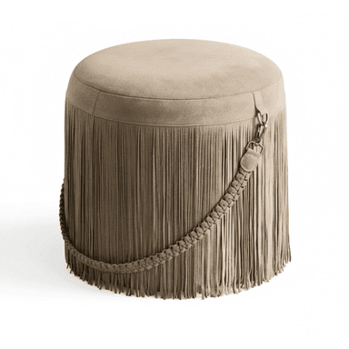 A cylindrical upholstered ottoman featuring a full skirt of cascading fringe around its perimeter. The piece is covered in a soft, matte taupe or greige suede-like material. A decorative braided strap, also in the same material, wraps diagonally around the body of the ottoman, secured by an antiqued brass-tone clasp. The top surface is flat and plush, indicating suitability for seating or footrest use. The overall aesthetic leans towards a bohemian or eclectic style. — 3D model (AI enhanced source)
