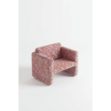 A contemporary, low-profile armchair featuring a distinctive multicolored, highly textured upholstery. The chair has a blocky, sculptural silhouette with integrated arms and backrest that flow seamlessly into the seat and base, giving it a continuous, monolithic appearance. The fabric presents a vibrant, speckled pattern composed of various hues including pink, red, orange, blue, purple, and yellow, creating a playful and eye-catching aesthetic. The base is also fully upholstered, eliminating visible legs. — 3D model (AI enhanced source)
