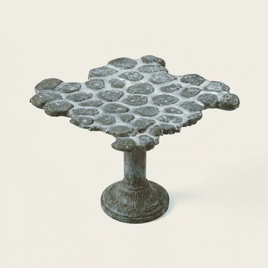 A unique pedestal table featuring an irregularly shaped tabletop composed of natural pebbles set in a white cementitious grout. The base is a single, ornate pedestal made of metal, likely bronze or copper, exhibiting a distinct verdigris patina, indicating age or a deliberate aged finish. The overall aesthetic is rustic and artisanal. — 3D model (AI enhanced source)