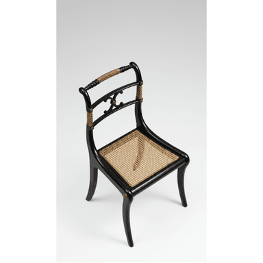 A refined Regency-style side chair featuring a black lacquered wooden frame, a woven cane seat, and decorative rope detailing on the back supports. The chair has a curved backrest with a carved central motif and subtly splayed saber legs. — 3D model (AI enhanced source)