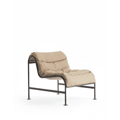 A contemporary lounge chair featuring a minimalist black metal frame that curves to support a deeply cushioned, upholstered seat and backrest. The frame is constructed from bent tubular metal, forming the legs and the continuous support for the textile cushions. The upholstery is a light beige or tan fabric, providing a soft contrast to the dark frame. The design is low-profile and appears designed for comfort and casual lounging. — 3D model (AI enhanced source)