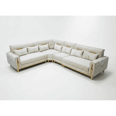 An L-shaped contemporary sectional sofa upholstered in a light cream or beige fabric, featuring distinct gold-toned metal accents on its arms and legs. The sofa has a low profile, plush seat cushions, and multiple decorative throw pillows. The backrests and sides exhibit channel tufting and diamond-patterned quilting in some areas, adding textural interest and a luxurious aesthetic. The overall design suggests a blend of modern sophistication and glam elements. — 3D model (AI enhanced source)