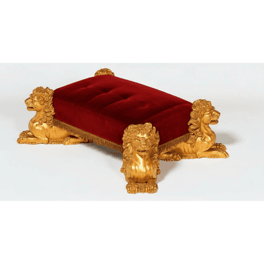 A rectangular ottoman featuring a deeply button-tufted red velvet upholstered top, supported by four intricately carved and gilded lion figures serving as legs. The upholstery is accented with a delicate gold fringe trim along its lower edge. — 3D model (AI enhanced source)