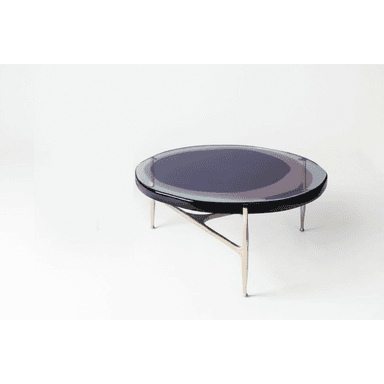 A round coffee table featuring a clear glass top with an iridescent finish that displays shifting colors, predominantly purple and blue tones. Beneath the glass is a thin black rim. The table is supported by a sculptural three-legged metal base with a warm, brass-like finish. The legs taper downwards and meet in a central point, giving the table a distinctive, modern appearance. — 3D model (AI enhanced source)