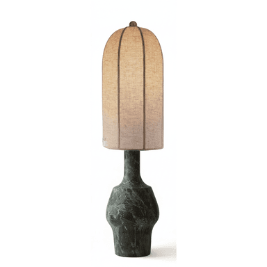 A distinctive table lamp featuring a heavily patinated, sculptural metal base in a dark green-bronze hue, supporting a tall, arched fabric shade. The base has an organic, fluid shape with a narrower neck and a wider, bulbous lower section, suggesting a lost-wax casting or hammered finish with natural oxidation. The shade is a light, neutral-toned linen or similar textile, with visible vertical ribs or seams, culminating in a rounded dome at the top. A small, brass-toned finial secures the shade. The lamp emits a warm, diffused light. — 3D model (AI enhanced source)