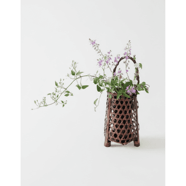 A traditional Japanese ikebana vase, meticulously crafted from dark, lacquered woven bamboo in a hexagonal pattern. It features a footed base for stability and an elegant arched handle. The vase holds a floral arrangement with delicate purple and white blossoms and trailing green foliage, presented against a stark white background. — 3D model (AI enhanced source)