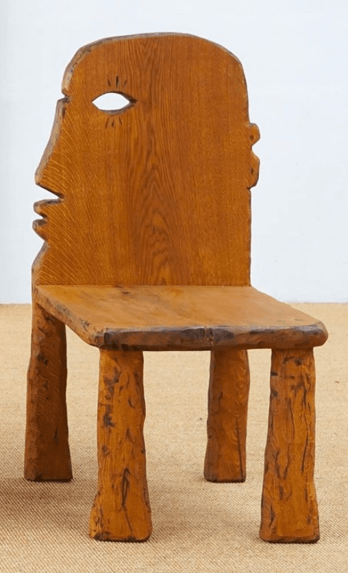 A distinctive carved wooden chair crafted from medium-brown oak, featuring an anthropomorphic backrest shaped like a human profile with a prominent eye cutout. The seat is a flat, solid square, supported by four chunky, irregularly shaped legs that contribute to its primitive, folk-art aesthetic. — 3D model