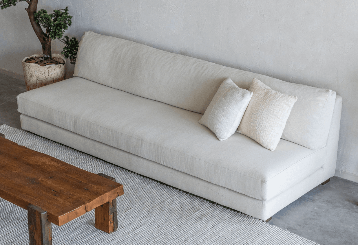 A modern, armless upholstered sofa in a light, textured fabric, featuring a low backrest and two matching throw pillows, supported by minimal wooden feet. — 3D model