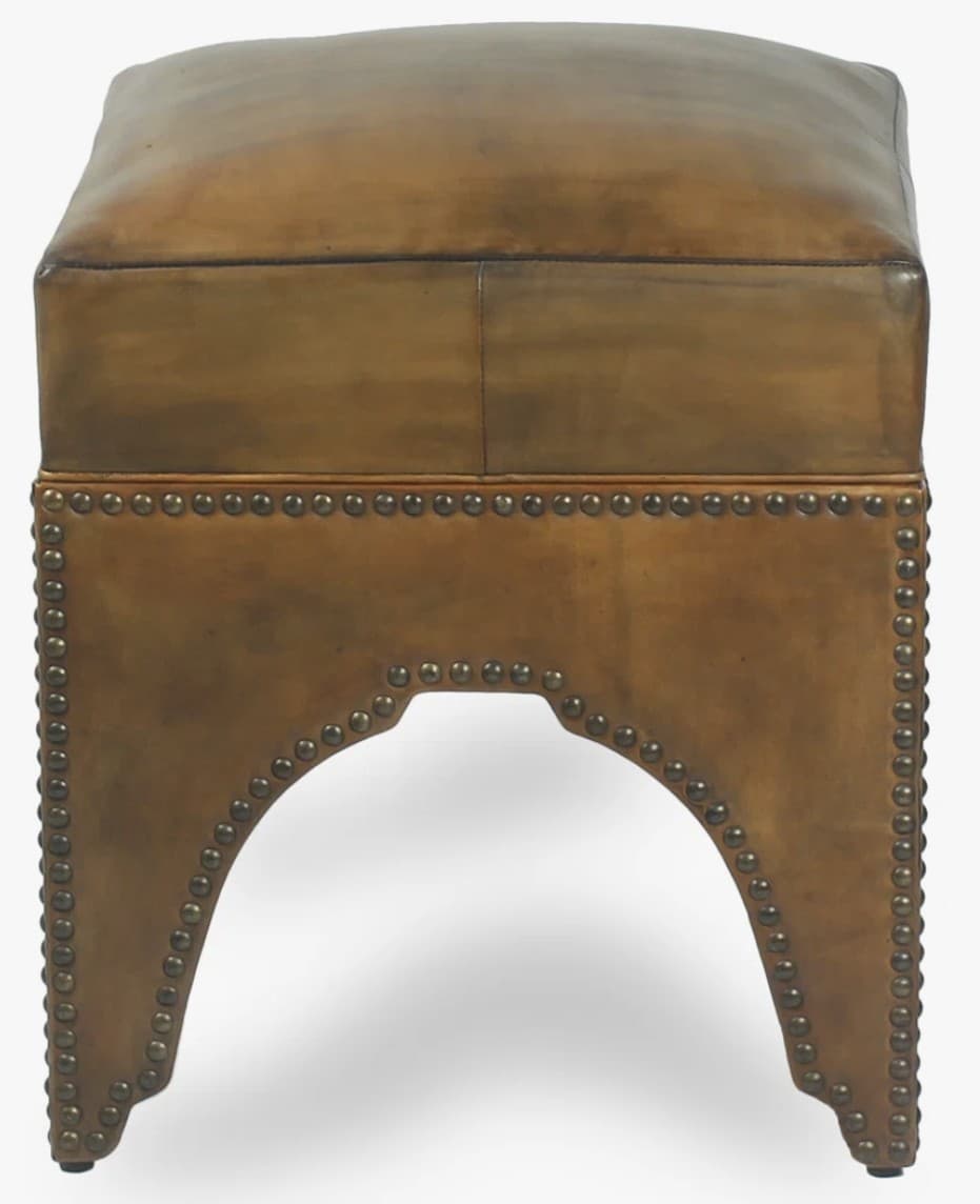 A square-shaped ottoman with a cushioned top, upholstered in what appears to be aged or distressed leather. The base is adorned with decorative brass or bronze nailhead trim. The sides of the base feature an arched cutout. — 3D model