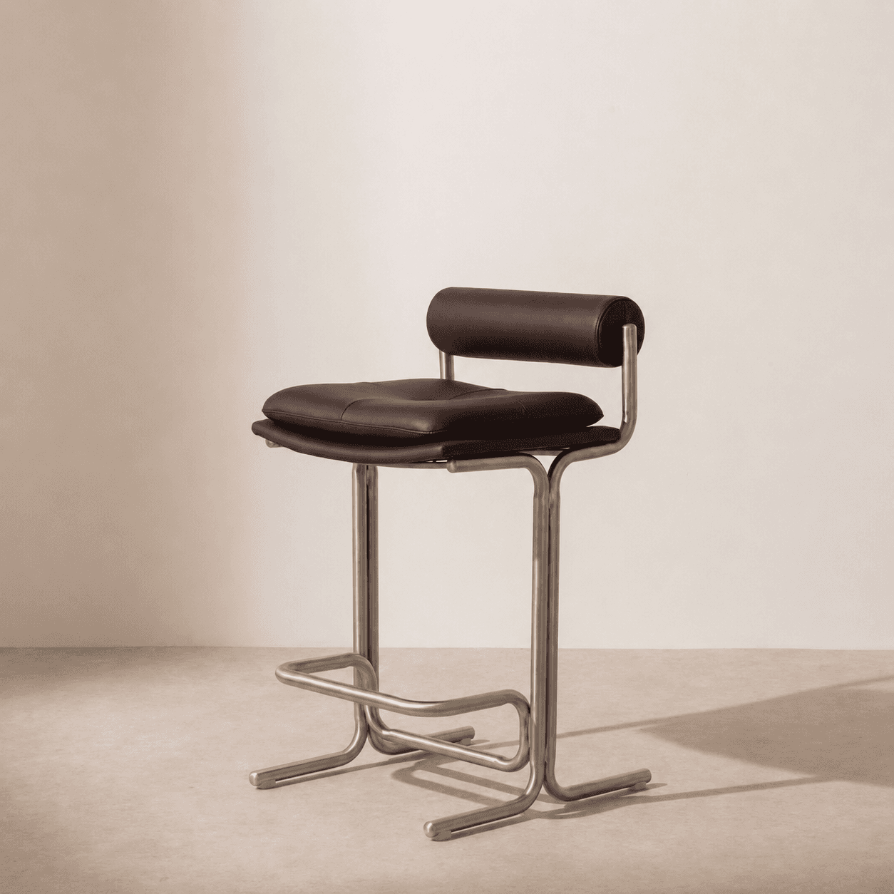A modern bar stool featuring a tubular metal frame with a brushed finish, brown leather upholstery on the seat, and a cylindrical backrest. The seat is padded and includes button-tufting or quilting details. An integrated footrest is part of the metal frame. — 3D model