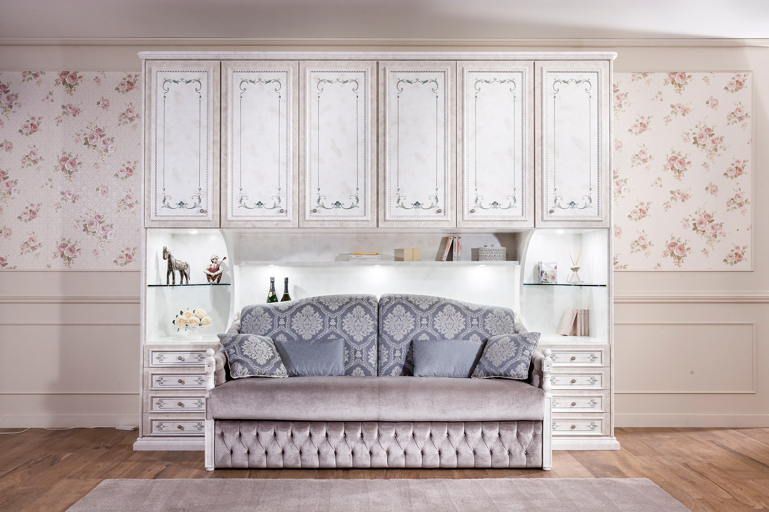 An ornate multifunctional wall unit featuring an integrated sofa bed, side storage, and upper cabinets. The sofa has a grey velvet upholstery with a tufted base and patterned back cushions. The surrounding cabinetry is finished in a light, possibly distressed or painted white wood, with decorative framed panels and subtle floral motifs. The unit also includes open display shelves with recessed lighting and two sets of drawers on either side of the sofa. — 3D model