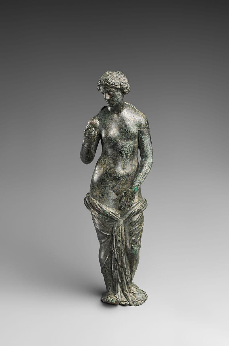 A standing classical bronze figurine depicting a female nude figure, likely Venus, with drapery artfully covering her lower body and modesty. Her head is tilted slightly downwards and to the left, and she holds a small, round object in her left hand. The bronze exhibits significant green and dark brown patination, indicating its age and material composition. The figure stands in a contrapposto pose, reflecting classical artistic conventions. — 3D model