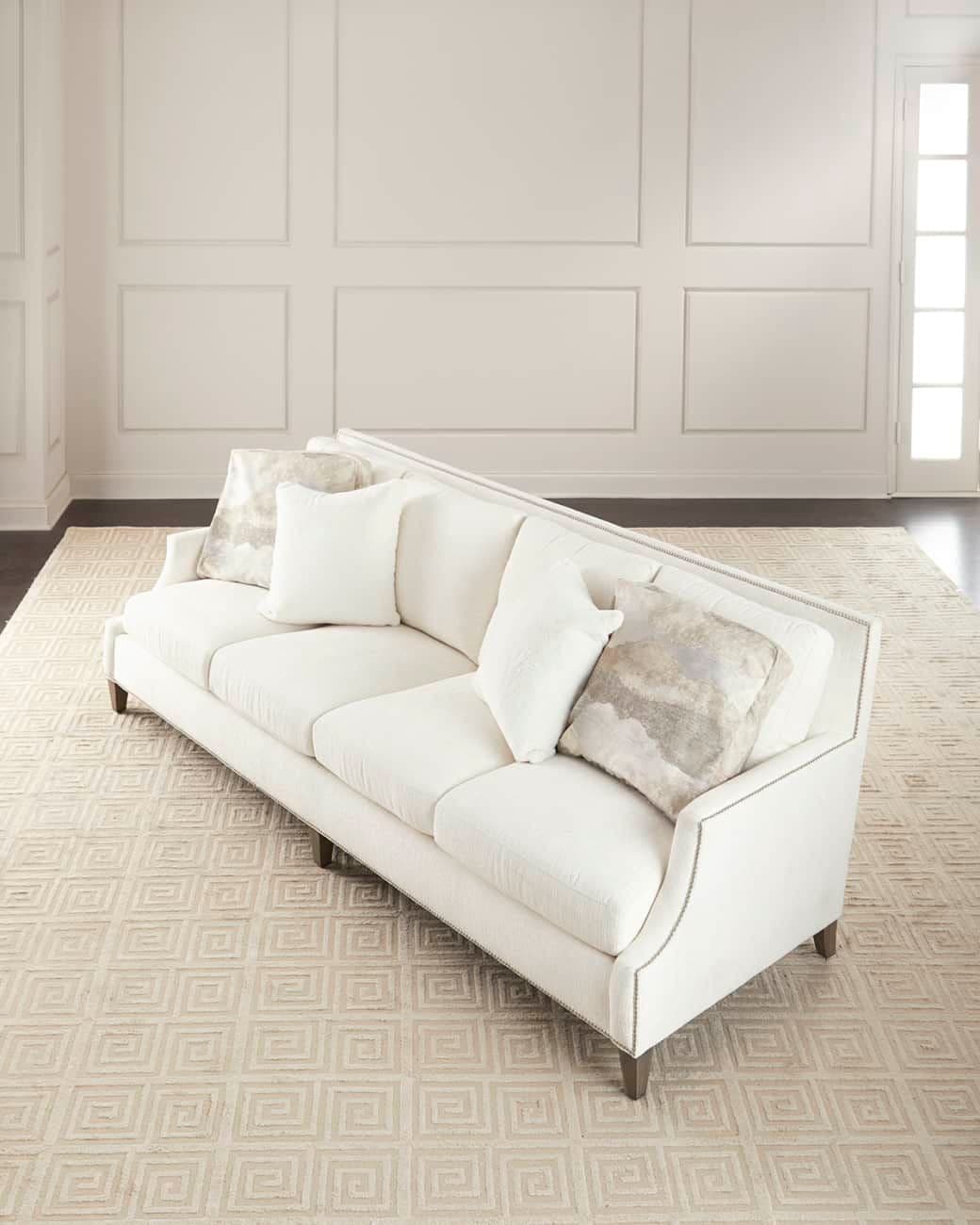 A white upholstered three-seater sofa with curved arms and decorative nailhead trim along the edges of the arms and back. It is accompanied by throw pillows, one of which has a marble-like pattern. The sofa is positioned on a cream-colored rug with a geometric pattern and rests on short, dark wooden legs. — 3D model