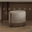 A cylindrical upholstered ottoman featuring a two-tone design. The upper body is covered in light beige fabric, while the lower section is wrapped in a contrasting brown leather. A matching brown leather handle is integrated into the side for portability. The top surface and edges are softly rounded, and the fabric sections feature piping detail. — 3D model