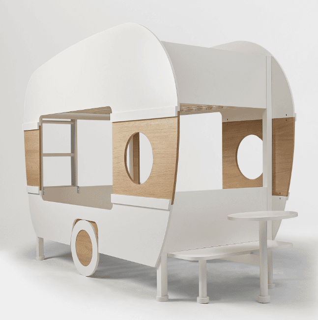 A children's bed designed in the whimsical shape of a caravan or camper, featuring a primary white finish with natural wood accents, cut-out windows, an integrated side table, and decorative wheels. — 3D model