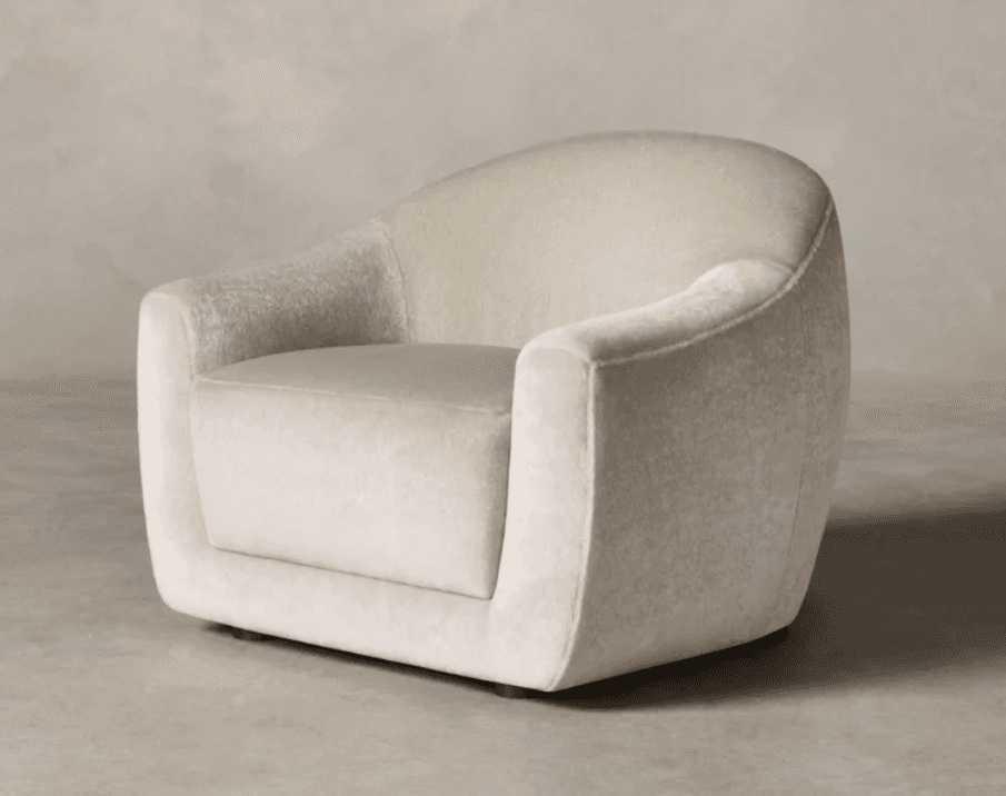 A contemporary club armchair upholstered in a light beige, textured fabric, resembling bouclé. The chair features a distinctive rounded, barrel-like silhouette with a wide, deep seat and integrated, subtly curved armrests that flow seamlessly into the backrest. The entire piece is generously padded, presenting a soft, inviting form. Small, dark wooden feet are visible at the base, slightly lifting the chair off the floor. Its design prioritizes comfort and a modern, organic aesthetic. — 3D model