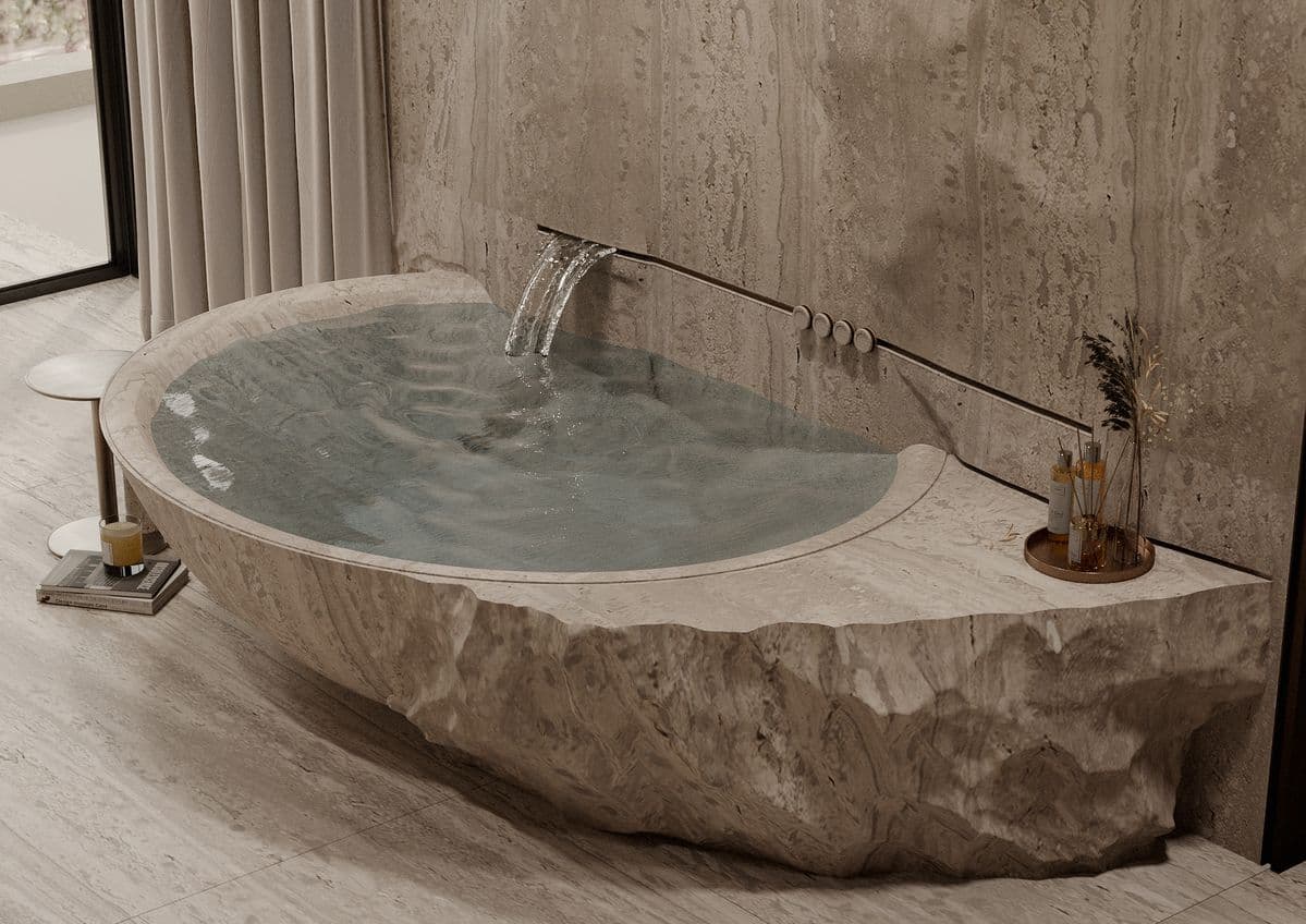 A freestanding bathtub carved from natural stone, with a rough, chiseled exterior resembling a rock formation. The interior is smooth and contains water. A waterfall faucet integrated into the stone wall behind the tub provides a gentle stream of water. The tub is partially recessed into a stone-tiled wall. — 3D model
