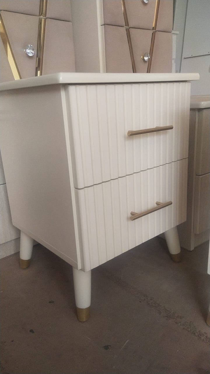 A modern two-drawer nightstand featuring a light neutral finish and elegant design details. The front of the drawers are adorned with vertical fluting, complemented by slender, horizontally oriented brass-toned bar pull handles. The nightstand stands on four tapered legs, each capped with a matching brass-toned accent at the bottom. — 3D model