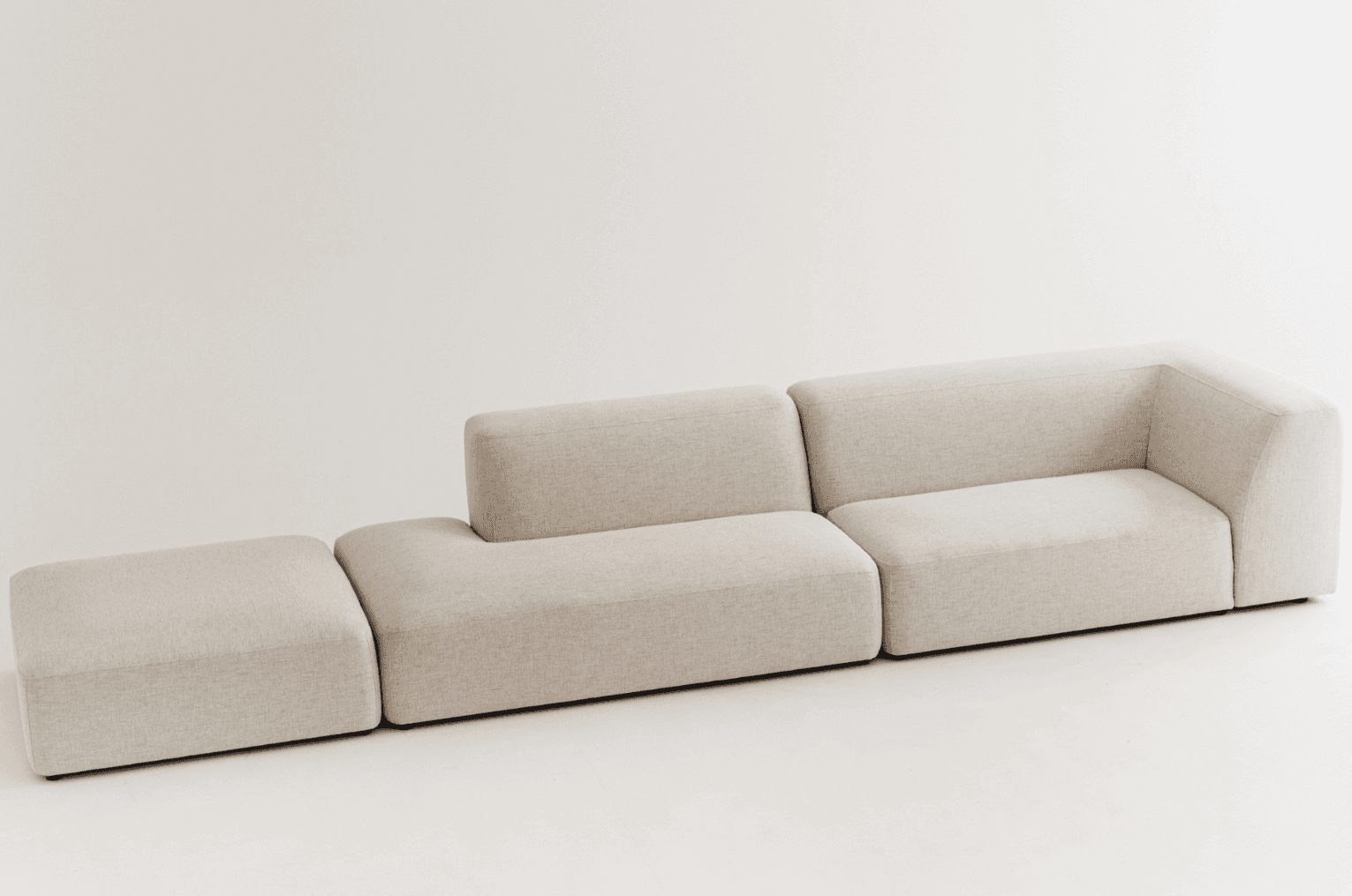 A light beige modular sectional sofa featuring three distinct components: a rectangular ottoman, a corner seating unit, and an armless seating unit. The design is contemporary with clean lines, thick block-like forms, and softly rounded edges. The upholstery is a textured woven fabric in an oatmeal or light beige hue. The arrangement suggests a flexible seating solution that can be reconfigured. — 3D model