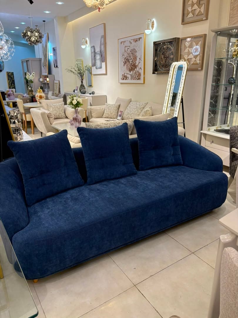 A modern three-seater sofa upholstered in a textured dark blue fabric. It features softly rounded armrests, a single long seat cushion, and three individual back cushions with button tufting. The sofa is supported by four small, light-colored wooden legs, visible at the corners. — 3D model