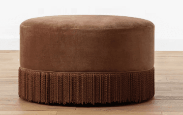 A contemporary round ottoman featuring plush velvet upholstery and a decorative fringe trim around its base. The piece has a solid, cylindrical form factor and is finished in a warm taupe-brown color. — 3D model
