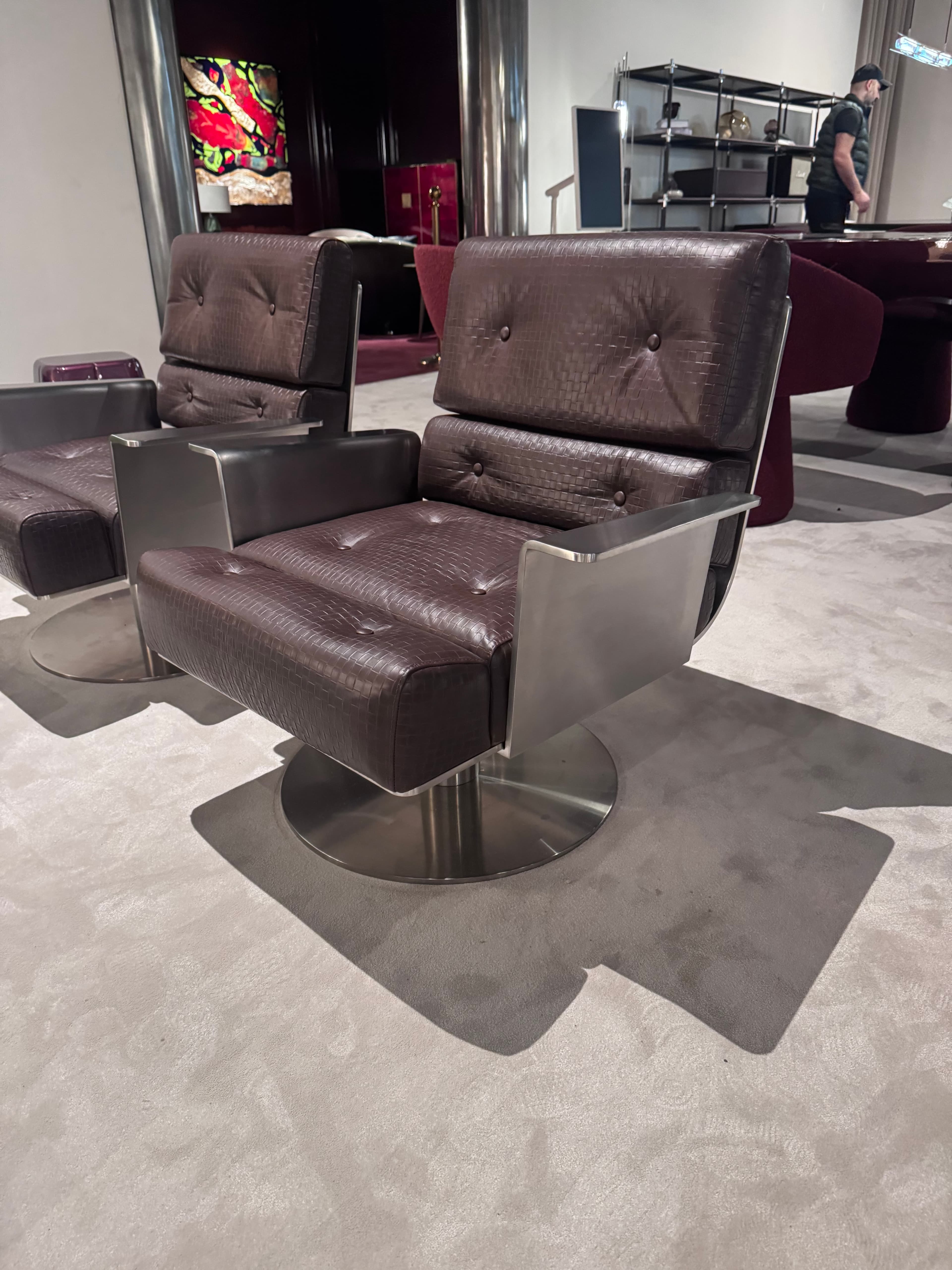 A modern armchair featuring a swiveling base. The seat and backrest are upholstered in dark brown leather with a textured, possibly embossed, finish that resembles crocodile skin. The upholstery is tufted with visible buttons. The armrests are integrated with the frame, which is constructed from brushed metal, offering a metallic contrast to the leather. The base is a circular, flat metal pedestal, suggesting a swivel mechanism for rotation. The overall design presents a blend of comfort and industrial aesthetic. — 3D model