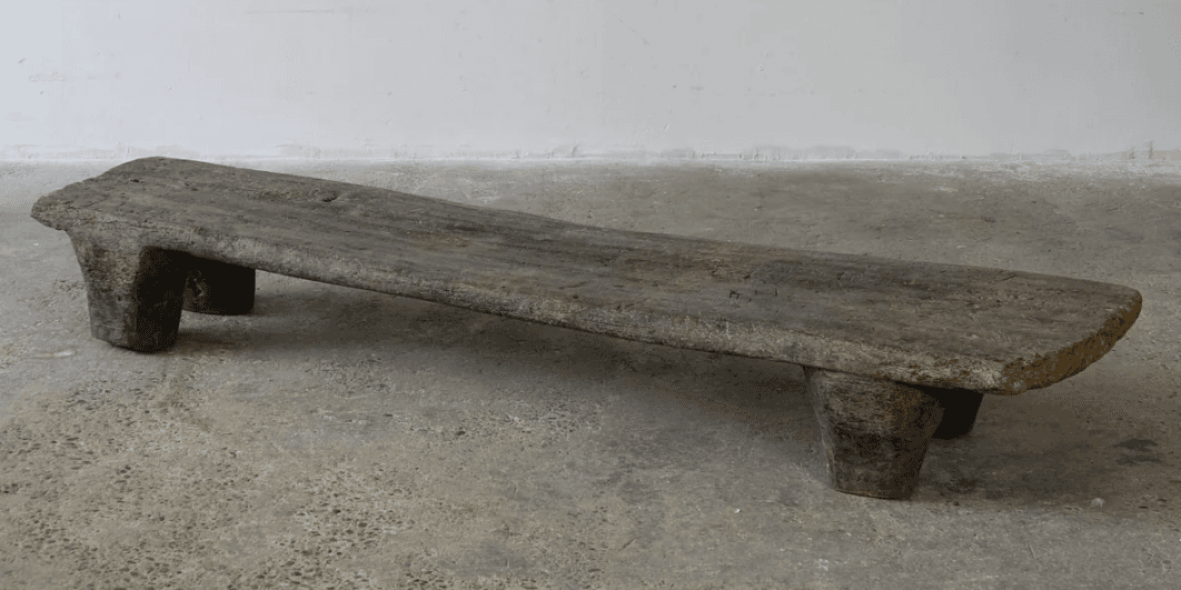 A rustic, low-profile bench with a flat, elongated top and two substantial, block-like legs at each end. The piece exhibits a weathered texture and irregular edges, suggesting it is made from natural wood. The design is simple and primitive. — 3D model