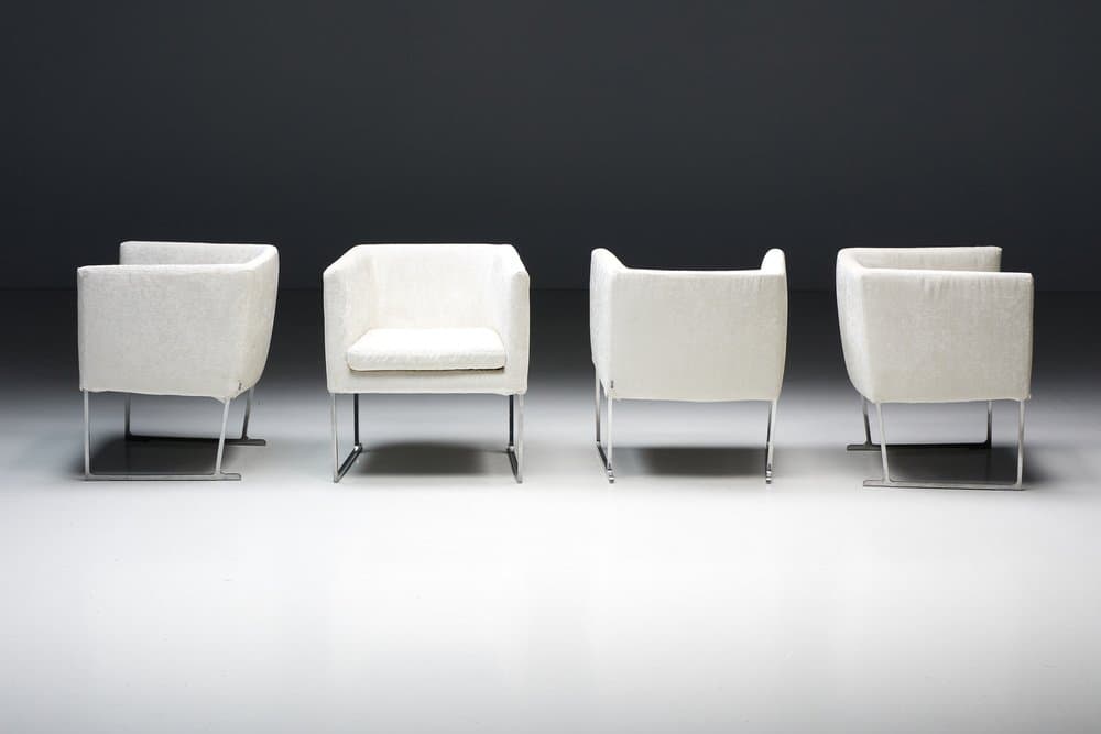 A set of four identical armchairs with a modern design. Each armchair features a cube-like upholstered seat and backrest with squared arms. The upholstery appears to be a textured fabric. The chairs are supported by a sleek, minimalist frame, likely made of polished metal, elevating them slightly off the ground. The overall aesthetic suggests a contemporary or minimalist style. — 3D model