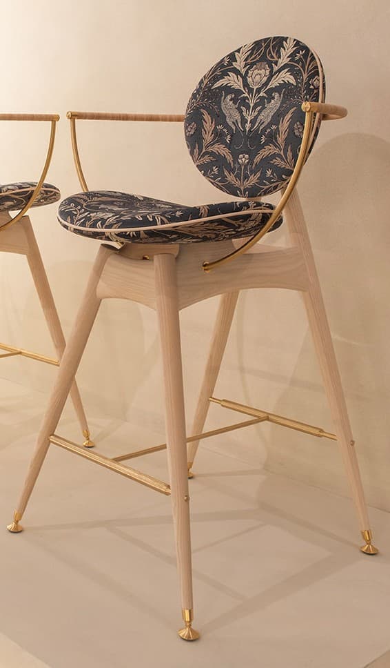 A tall, contemporary bar stool featuring a light-toned wooden frame, brass accents, and distinctive dark, patterned upholstery. The seat and backrest are circular and upholstered with a black fabric featuring an intricate floral and animal pattern in gold and bronze tones. The backrest is supported by curved brass elements that also form integrated armrests. The splayed wooden legs are reinforced by brass stretchers that serve as a footrest and terminate in brass caps. — 3D model