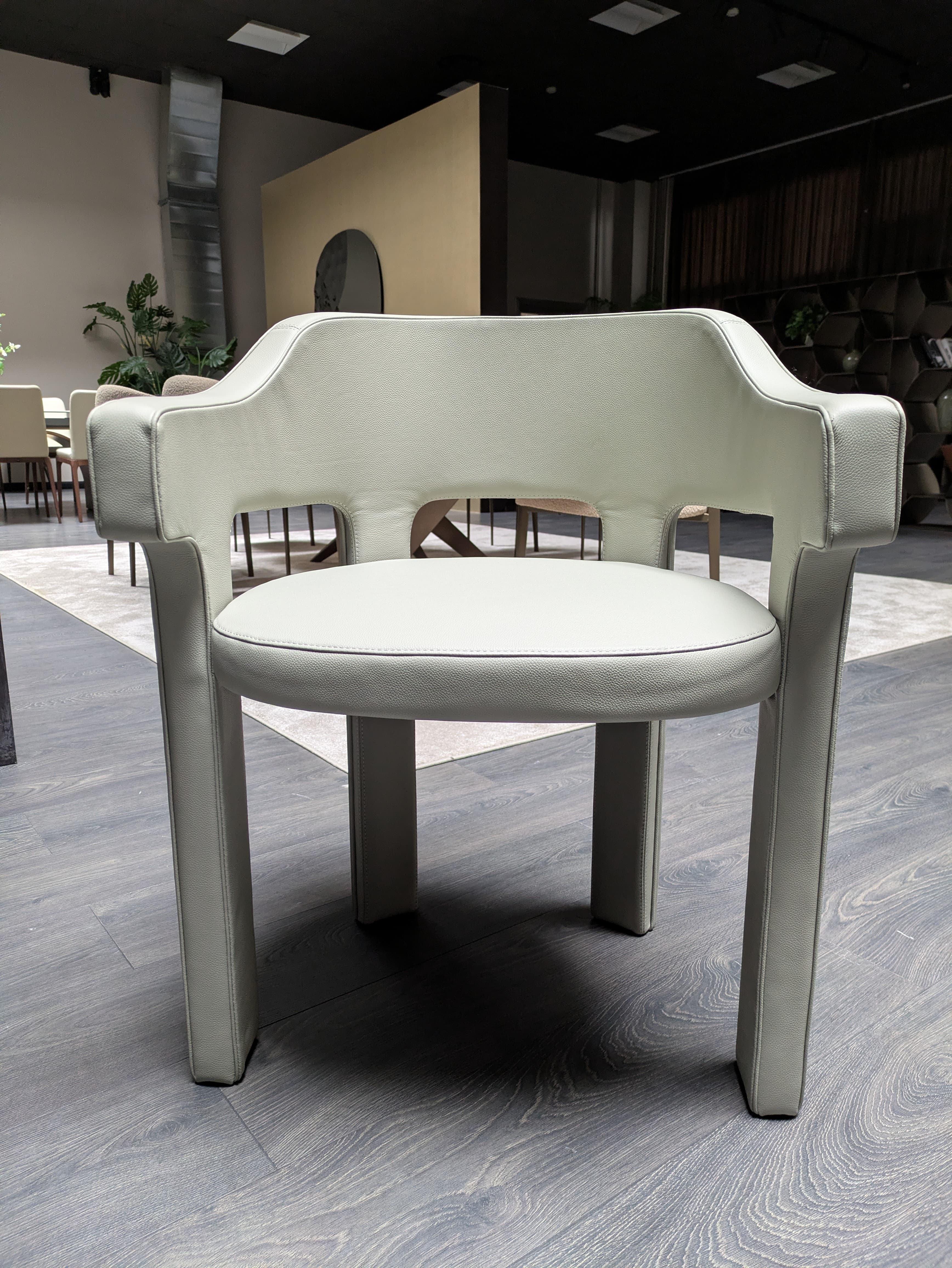 A modern armchair fully upholstered in light grey, pebbled faux leather or leather. It features a distinctive design with arms seamlessly integrated into the backrest, creating a continuous, rounded form. The seat is cushioned and the chair stands on four substantial, upholstered legs, contributing to its monolithic and elegant aesthetic. The overall impression is one of minimalist luxury and comfort. — 3D model