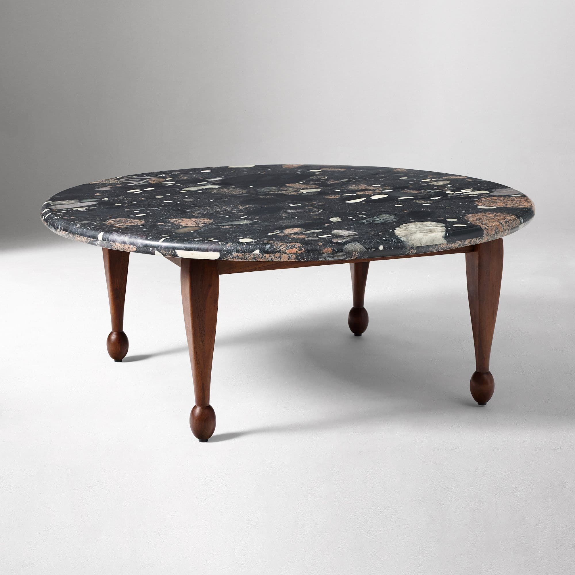 An oval-shaped coffee table with a dark, speckled stone top and four wooden legs. The tabletop features a mosaic of various stone fragments, predominantly dark gray, with inclusions of lighter gray, white, beige, and reddish-brown pieces. The legs are tapered and appear to be made of dark wood, with a rounded knob at the base of each. — 3D model