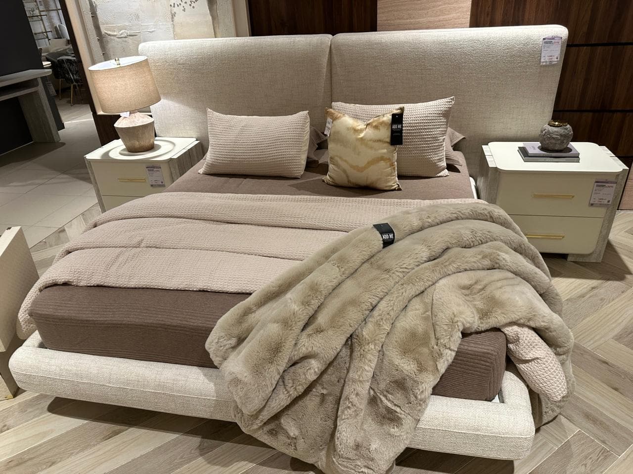 An upholstered platform bed featuring a clean, minimalist design with a distinctive two-panel headboard and an integrated fabric-wrapped frame. The bed is presented with neutral-toned bedding, including a brown fitted sheet, a light beige knit blanket, and a faux fur throw, complemented by various decorative pillows. — 3D model