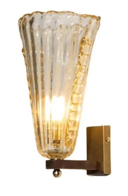 A wall-mounted light fixture featuring an amber-tinted, ribbed glass shade that flares outwards towards the top, resembling a trumpet or cornucopia. The shade has a textured, fluted surface and appears to be artisanal with a slightly irregular, scalloped upper edge. It is supported by a simple brass-toned metal arm and wall plate, designed to cast a warm, diffused light. The light source is visible within the glass shade, indicating it is an active fixture. — 3D model