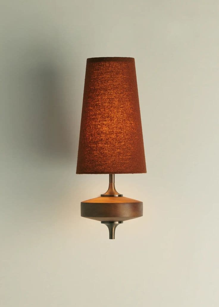 A wall-mounted lamp with a conical lampshade and a distinct, multi-tiered base. The lampshade appears to be made of textured fabric, possibly linen or burlap, in a warm, reddish-brown hue. The base is constructed from wood, featuring a wider, flattened circular element with a concave underside, mounted on a metallic stem. The overall design suggests a mid-century modern or retro aesthetic. — 3D model