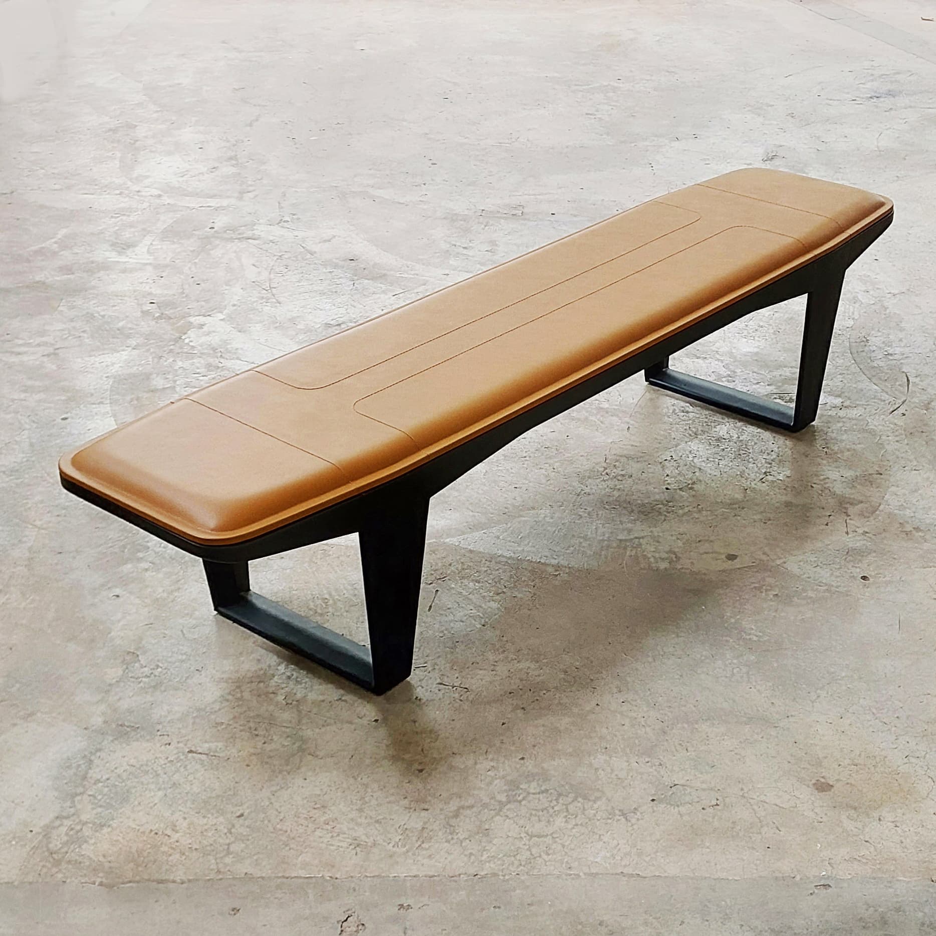 A long, rectangular bench featuring a padded, tan upholstered seat with stitched detailing, supported by a minimalist black metal frame with inverted V-shaped legs. — 3D model