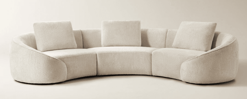 A curved sectional sofa with a textured upholstery and plush throw pillows. The sofa features a low profile and rounded arms, offering a modern, organic aesthetic. Its modular design suggests adaptability to various spatial configurations. — 3D model