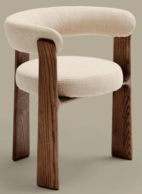 A modern armchair with a curved upholstered backrest and seat, supported by four solid wood legs. The legs have a distinct organic shape and visible wood grain. The upholstery is a textured, light-colored fabric. The design is minimalist and contemporary. — 3D model