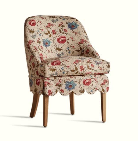 An armchair with a floral fabric upholstery. It features a high back that curves gently to the sides, a padded seat cushion, and four wooden legs. The base of the chair has a decorative scalloped trim. The fabric pattern consists of a cream background with scattered red and blue flowers, green leaves, and small dots. — 3D model