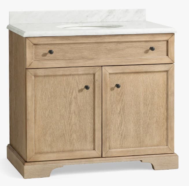 A freestanding bathroom vanity with a marble top featuring an integrated sink basin. The base unit is constructed from wood with a light, natural finish, exhibiting a prominent wood grain pattern. It comprises a single drawer above two cabinet doors, each with a dark, round knob. The base of the vanity has a decorative plinth. — 3D model