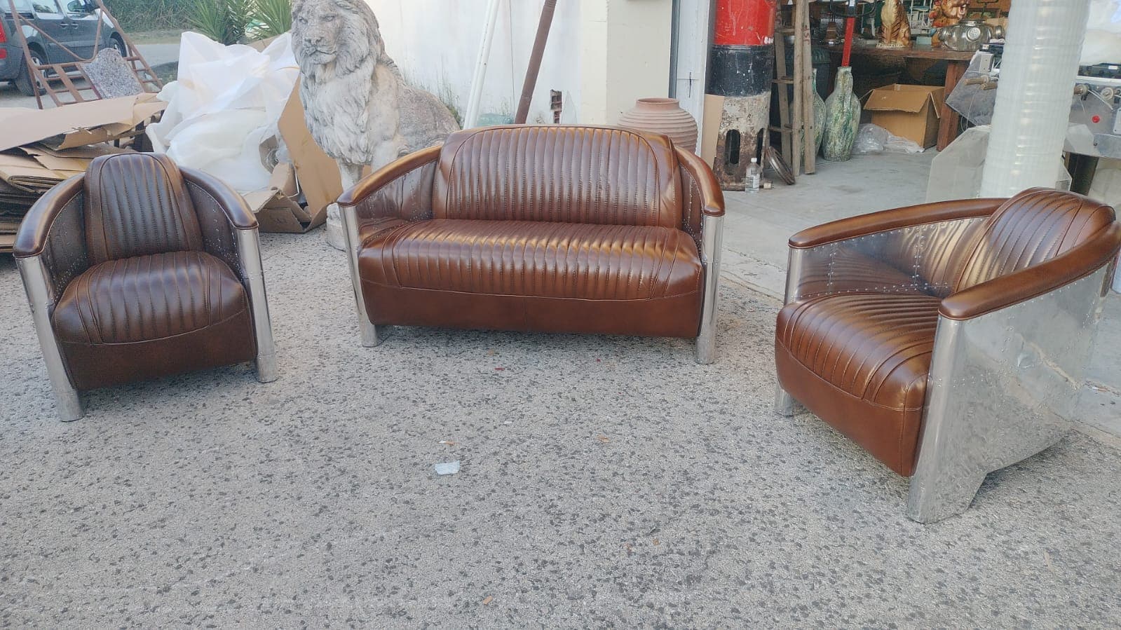 A two-seater sofa with a distinct aviator-inspired design. The seating and backrest are upholstered in brown, tufted leather, featuring vertical channel stitching. The arms and the outer sides of the sofa are clad in brushed aluminum panels, riveted along the edges, resembling aircraft fuselage construction. The sofa has a slightly curved silhouette and sits on low-profile, angled metal legs. — 3D model