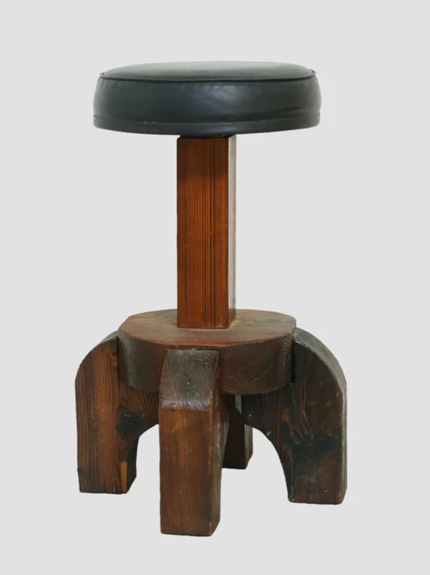 A robust, brutalist-style stool featuring a circular, dark upholstered seat atop a thick, central wooden column. The base is constructed from heavy, blocky pieces of dark-stained wood, forming a sturdy, four-legged support structure with a distinct, architectural aesthetic. — 3D model