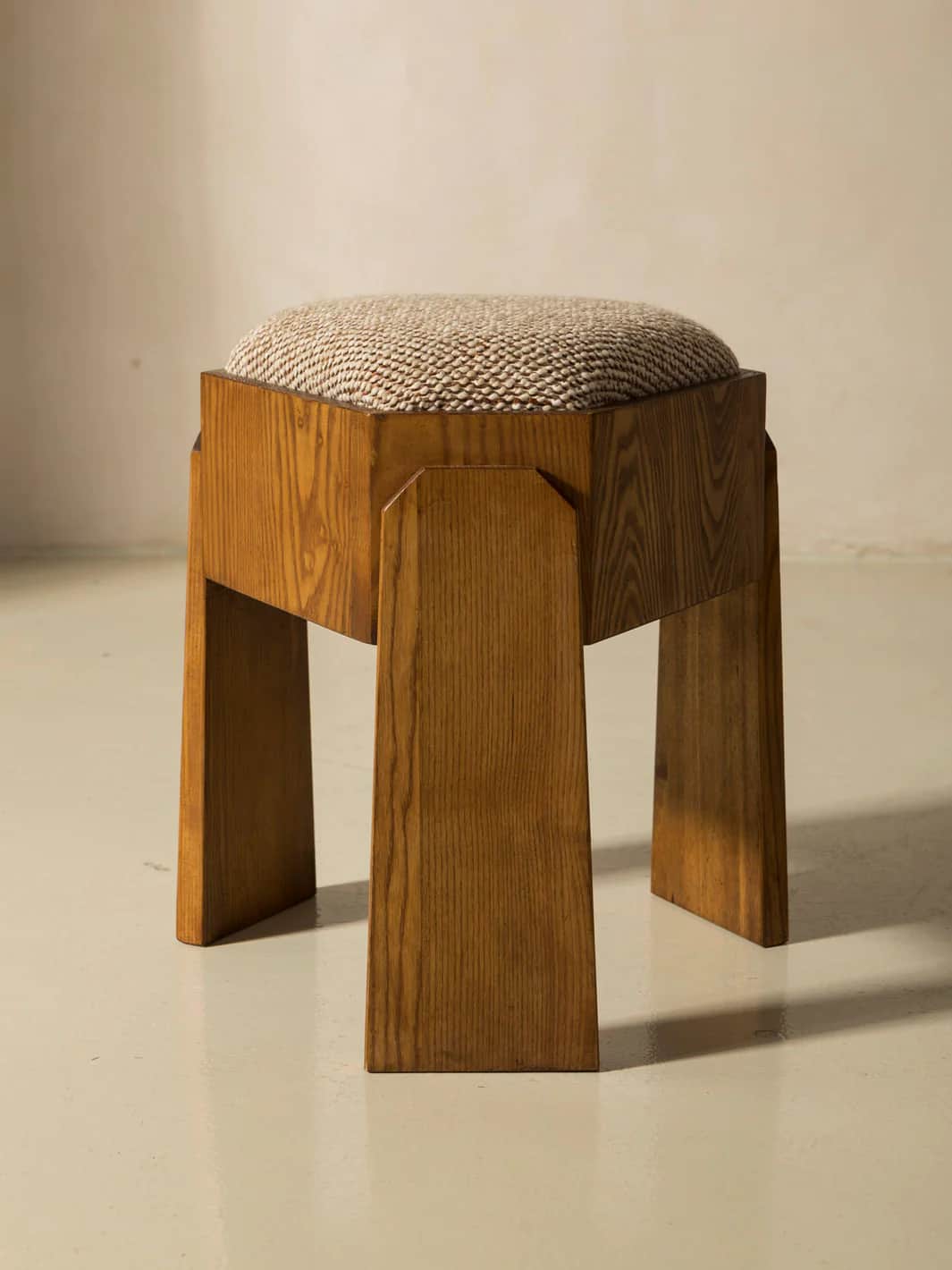 A wooden stool with a padded top. The stool features four legs with a geometric design, tapering sli… — 3D model