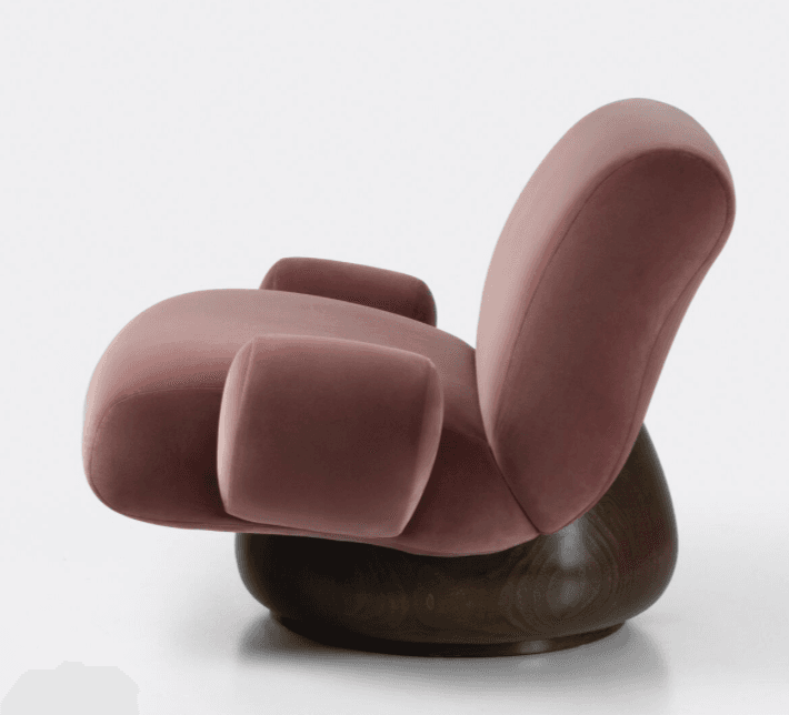A plush armchair with a rounded silhouette and voluminous cushioning. The seat and backrest are upholstered in a soft, rose-colored velvet fabric. It features integrated armrests that flow seamlessly from the main body of the chair. The chair is supported by a substantial, dark-stained wooden base with a rounded, drum-like shape. — 3D model