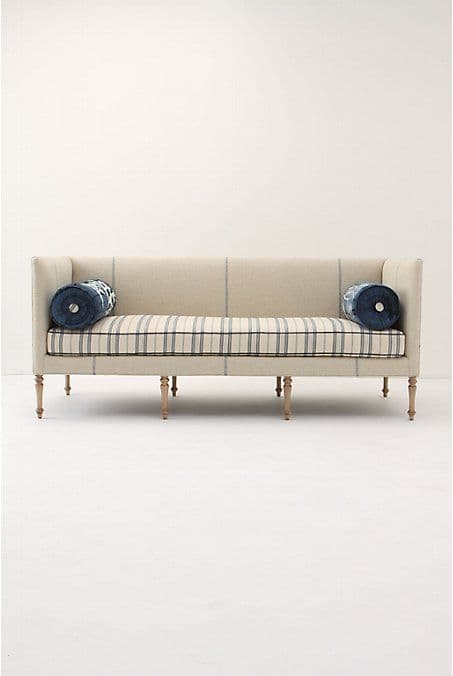 An upholstered daybed settee featuring a beige fabric with a subtle plaid pattern on the exterior frame and a blue and white striped pattern on the seat cushion. It has a single high backrest and a low armrest on the opposite side, creating an open, elongated seating area. The settee is supported by six light-colored, turned wooden legs. Two round bolster pillows, upholstered in dark blue fabric with white embroidered patterns, are positioned at each end. — 3D model