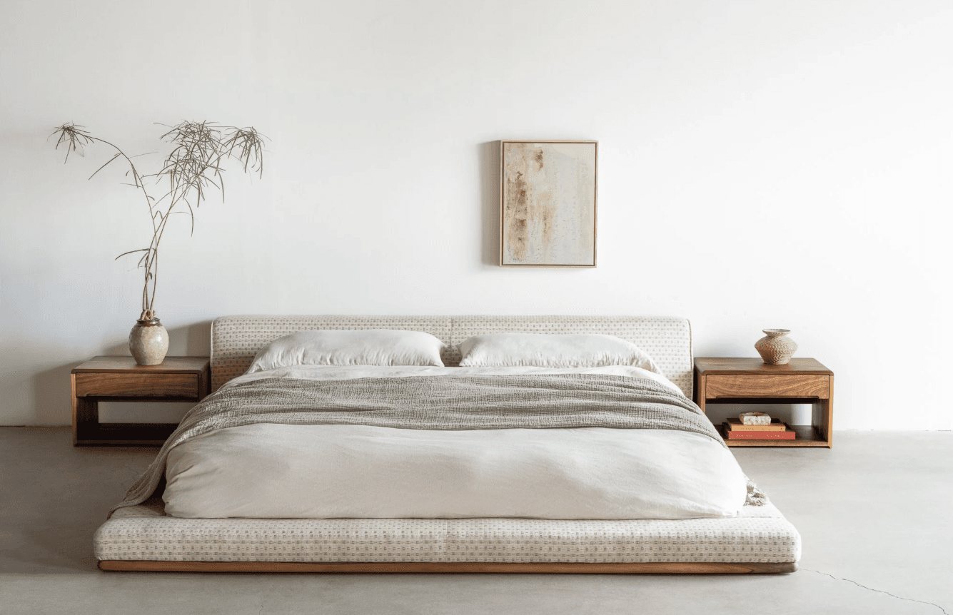 A low-profile platform bed with a textured mattress base and a white duvet with two pillows and a grey throw blanket. Two floating wooden nightstands are attached to the sides of the bed frame, each with a small vase or decorative object and books on the right nightstand. — 3D model